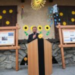 Ein Mann spricht auf einem Podium vor einer dekorierten Wand mit Sonnenblumen, Wolken und Informationsplakaten auf Holzbrettern.