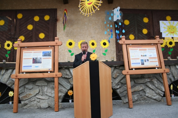 Ein Mann spricht auf einem Podium vor einer dekorierten Wand mit Sonnenblumen, Wolken und Informationsplakaten auf Holzbrettern.