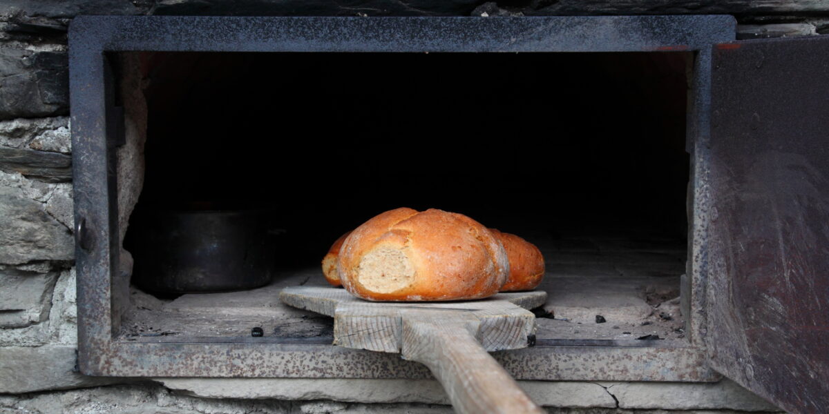 Ein Laib Brot auf einem Holzbrett wird in einen Steinofen mit einer Metalltür gelegt oder aus diesem herausgenommen.