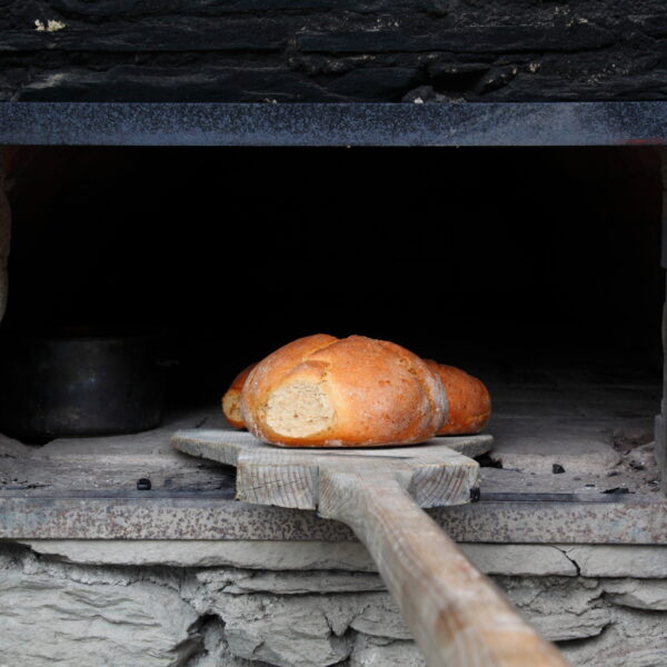Ein Laib Brot auf einem Holzbrett wird in einen Steinofen mit einer Metalltür gelegt oder aus diesem herausgenommen.