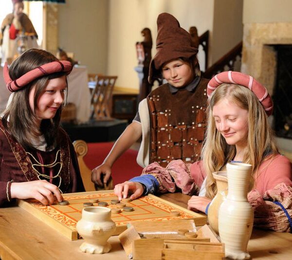 Drei Personen in mittelalterlichen Kostümen spielen ein historisches Brettspiel an einem Holztisch, mit Töpferwaren und einer Feuerstelle im Hintergrund.
