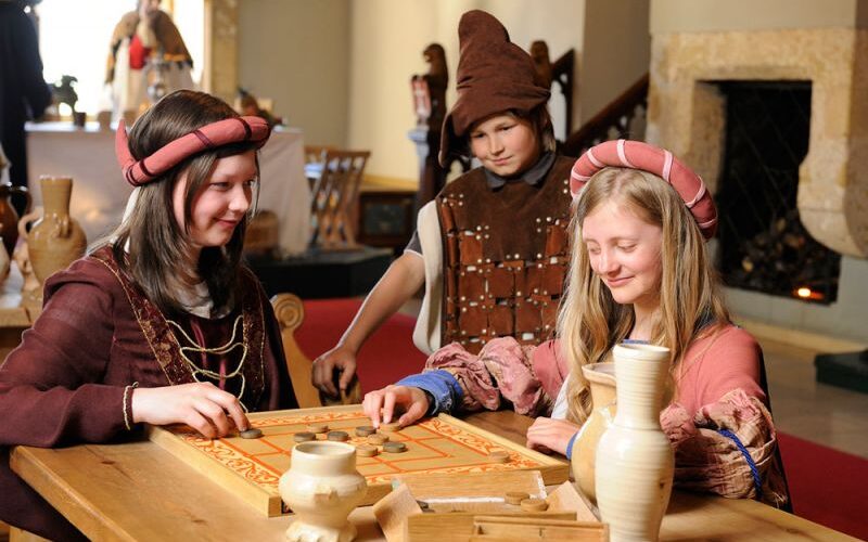 Drei Personen in mittelalterlichen Kostümen spielen ein historisches Brettspiel an einem Holztisch, mit Töpferwaren und einer Feuerstelle im Hintergrund.