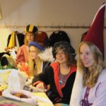 Vier Personen sitzen an einem Tisch und tragen mittelalterliche Kostüme und Hüte, während im Hintergrund weitere Kostüme an Haken hängen.