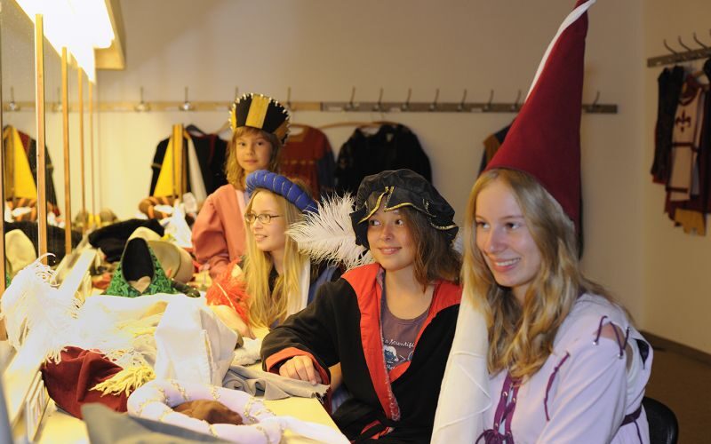 Vier Personen sitzen an einem Tisch und tragen mittelalterliche Kostüme und Hüte, während im Hintergrund weitere Kostüme an Haken hängen.