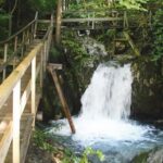 Ein kleiner Wasserfall mündet in ein felsiges, von Grün umgebenes Becken, über das ein Holzsteg und ein Fußweg führen.