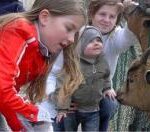 Drei Kinder streicheln und beobachten eine Ziege durch einen Zaun in einem Außenbereich.