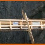 Eine hölzerne Fußgängerbrücke mit Geländer überspannt eine felsige Schlucht, auf der mehrere Personen entlanggehen.