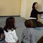 Eine Frau sitzt auf dem Boden und unterrichtet eine Gruppe kleiner Kinder an einer Tafel mit Klaviertasten und Musiknoten.