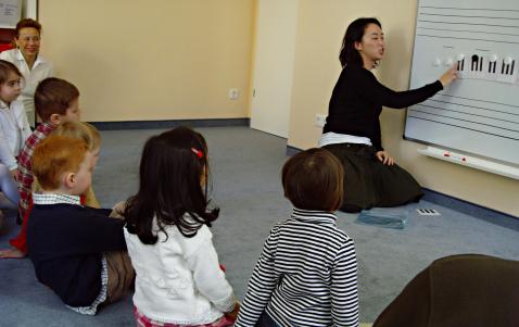 Eine Frau sitzt auf dem Boden und unterrichtet eine Gruppe kleiner Kinder an einer Tafel mit Klaviertasten und Musiknoten.