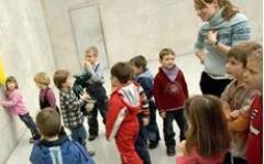 Eine Gruppe kleiner Kinder steht in einem Raum mit einer erwachsenen Frau, einige stehen mit dem Gesicht zur Wand, andere unterhalten sich oder warten.