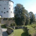 Ein runder weißer Steinturm steht auf einem grasbewachsenen Hügel, der von Steinmauern und Bäumen umgeben ist, während im Hintergrund die Berge zu sehen sind.