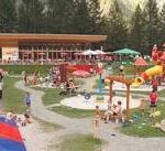 Kinder und Erwachsene treffen sich auf einem Spielplatz im Freien und einem Erholungsgebiet mit Spielgeräten, Sand und Picknicktischen, umgeben von Bäumen und Bergen.