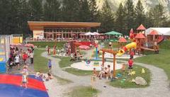 Kinder und Erwachsene treffen sich auf einem Spielplatz im Freien und einem Erholungsgebiet mit Spielgeräten, Sand und Picknicktischen, umgeben von Bäumen und Bergen.