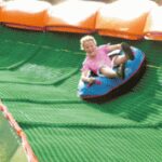 Eine Person fährt mit einem blauen aufblasbaren Schlauch eine grüne künstliche Rutsche hinunter, umgeben von orangefarbenen Polstern.