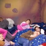 In einem Raum mit bemalten Steinwänden liegen und faulenzen mehrere Kinder auf einem Stapel von blauen Kissen.