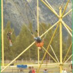 Zwei Personen hüpfen auf Trampolinen mit Gurten und Bungee-Seilen, umgeben von einem gelben Metallrahmen in einer Parklandschaft.