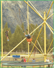Zwei Personen hüpfen auf Trampolinen mit Gurten und Bungee-Seilen, umgeben von einem gelben Metallrahmen in einer Parklandschaft.