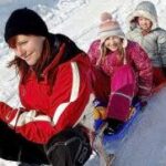 Ein Erwachsener und zwei Kinder in Winterkleidung sitzen gemeinsam auf einem Schlitten und rutschen lächelnd einen verschneiten Hügel hinunter.