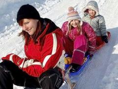 Ein Erwachsener und zwei Kinder in Winterkleidung sitzen gemeinsam auf einem Schlitten und rutschen lächelnd einen verschneiten Hügel hinunter.