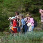 Eine Gruppe von Kindern und ein Erwachsener stehen dicht beieinander am Rande eines Teiches und blicken auf das Wasser, umgeben von grüner Vegetation und Bäumen.
