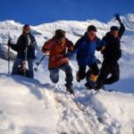 Vier Personen in Winterkleidung springen gemeinsam über einen verschneiten Berghang, mit schneebedeckten Gipfeln und blauem Himmel im Hintergrund.