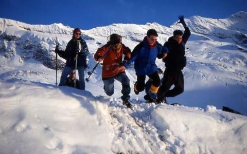 Vier Personen in Winterkleidung springen gemeinsam über einen verschneiten Berghang, mit schneebedeckten Gipfeln und blauem Himmel im Hintergrund.