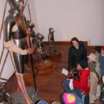 Eine Gruppe von Kindern und ein Erwachsener betrachten in einem Museum eine Ausstellung mit mittelalterlichen Rüstungen.