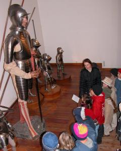 Eine Gruppe von Kindern und ein Erwachsener betrachten in einem Museum eine Ausstellung mit mittelalterlichen Rüstungen.