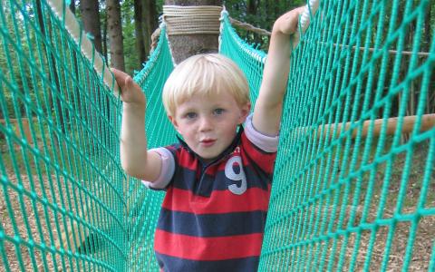 Ein Junge mit blondem Haar steht auf einer Seilbrücke im Freien und hält sich mit beiden Händen an dem grünen Netz fest.