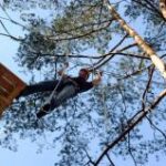 Eine Person in einem Gurtzeug seilt sich zwischen hölzernen Plattformen hoch oben in den Bäumen gegen einen blauen Himmel ab.