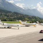 Mehrere Privatjets sind auf dem Rollfeld eines Flughafens geparkt, im Hintergrund sind Berge und eine Stadt zu sehen.