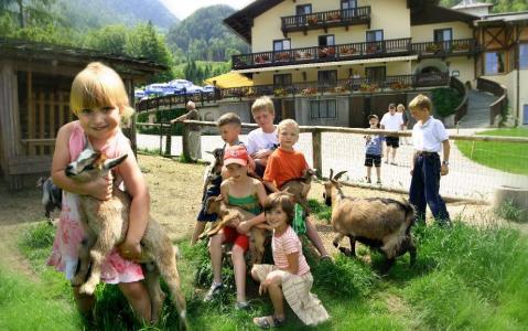 Mehrere Kinder spielen mit Ziegen auf einer Wiese in der Nähe eines Holzzauns, mit einem großen Gebäude und Bergen im Hintergrund.