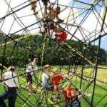 Kinder klettern auf einem geometrischen Klettergerüst im Freien auf einer Wiese, mit Bäumen im Hintergrund.