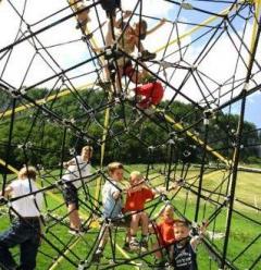 Kinder klettern auf einem geometrischen Klettergerüst im Freien auf einer Wiese, mit Bäumen im Hintergrund.
