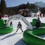 Kinder spielen auf einem verschneiten Hügel unter einem aufblasbaren Bogen mit der Aufschrift "Nici Bear Land Jungholz", während im Hintergrund Erwachsene und ein Skilift zu sehen sind.