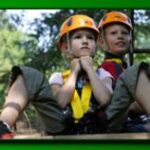 Zwei Kinder mit Helmen und Klettergurten sitzen im Freien und nehmen möglicherweise an einem Hochseilgarten oder einer Abenteueraktivität teil. Im Hintergrund sind Bäume und Grünflächen zu sehen.