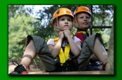 Zwei Kinder mit Helmen und Klettergurten sitzen im Freien und nehmen möglicherweise an einem Hochseilgarten oder einer Abenteueraktivität teil. Im Hintergrund sind Bäume und Grünflächen zu sehen.