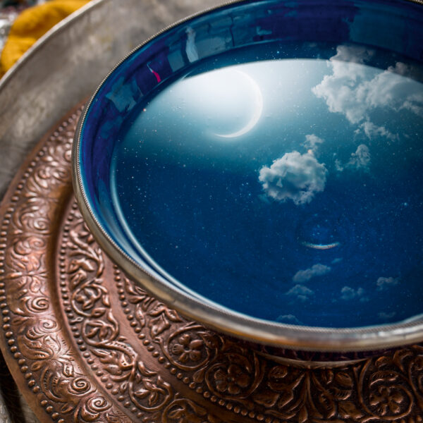 In einer blauen Schale mit Wasser auf einem verzierten Metalltablett spiegeln sich eine Mondsichel und Wolken, so dass in der Schale die Illusion eines Nachthimmels entsteht.