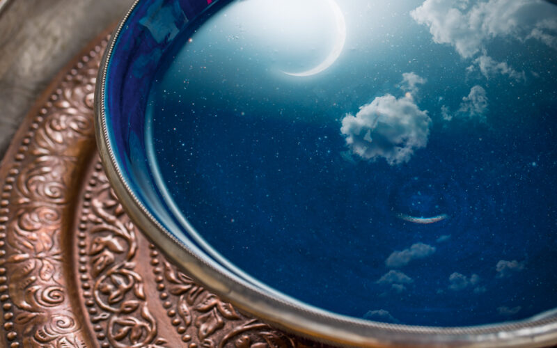 In einer blauen Schale mit Wasser auf einem verzierten Metalltablett spiegeln sich eine Mondsichel und Wolken, so dass in der Schale die Illusion eines Nachthimmels entsteht.