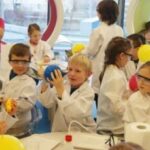 Kinder, die Laborkittel und Schutzbrillen tragen, nehmen an einer wissenschaftlichen Aktivität im Haus teil und hantieren mit Luftballons und Laborgeräten.