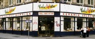 Straßeneckenansicht eines Keramikgeschäfts mit großen Fenstern und einem Schild mit der Aufschrift "Malen XL Keramik Selber Bemalen". Zwei Personen stehen in der Nähe des Eingangs.