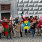 Eine Gruppe von Kindern posiert im Freien in Winterkleidung, mit bunten Pappschilden und Schwertern in der Hand, einige tragen Kegelhüte, vor einer gemusterten Wand.