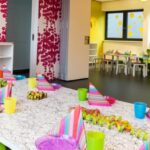 Ein bunt dekorierter Partyraum mit einem langen Tisch, der mit bunten Tellern, Tassen und Servietten gedeckt ist, kindgerechten Möbeln und verspielten rosa, gelben und weißen Wandmustern.