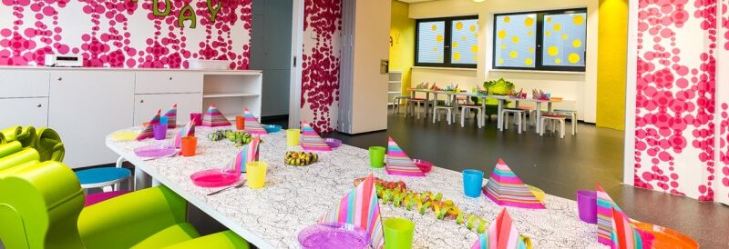 Ein bunt dekorierter Partyraum mit einem langen Tisch, der mit bunten Tellern, Tassen und Servietten gedeckt ist, kindgerechten Möbeln und verspielten rosa, gelben und weißen Wandmustern.