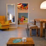 Ein gemütliches Zimmer mit einem gelben Sessel, Holztisch und -bänken, einer großen runden Lampe und einem Blick durch eine offene Tür in einen bunten Spielbereich.