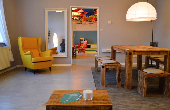 Ein gemütliches Zimmer mit einem gelben Sessel, Holztisch und -bänken, einer großen runden Lampe und einem Blick durch eine offene Tür in einen bunten Spielbereich.