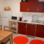 Kleine Küche mit roten Schränken, einer Spüle, einer Mikrowelle, einem Kühlschrank, einem Herd, einem Holztisch mit Stühlen und zwei roten Teppichen auf dem Boden.