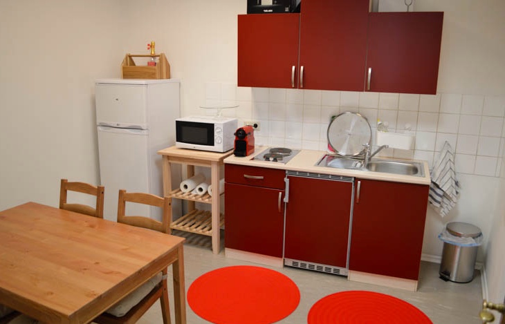 Kleine Küche mit roten Schränken, einer Spüle, einer Mikrowelle, einem Kühlschrank, einem Herd, einem Holztisch mit Stühlen und zwei roten Teppichen auf dem Boden.