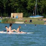 Drei Personen sitzen auf einem Paddelbrett auf einem See, andere entspannen sich am grasbewachsenen Ufer und ein Segelboot ist im Hintergrund zu sehen.