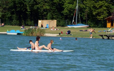 Drei Personen sitzen auf einem Paddelbrett auf einem See, andere entspannen sich am grasbewachsenen Ufer und ein Segelboot ist im Hintergrund zu sehen.